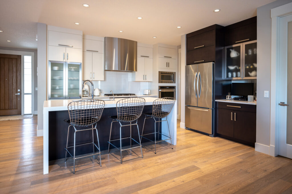 Modern kitchen with a large island, three wire bar stools, stainless steel appliances, white and dark wood cabinetry, built-in oven and microwave, and light wood flooring. Bright and spacious with recessed lighting.